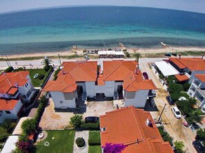An aerial view of a coastal residential area with houses featuring red-tiled roofs, a beach in the background, and a clear view of the ocean.