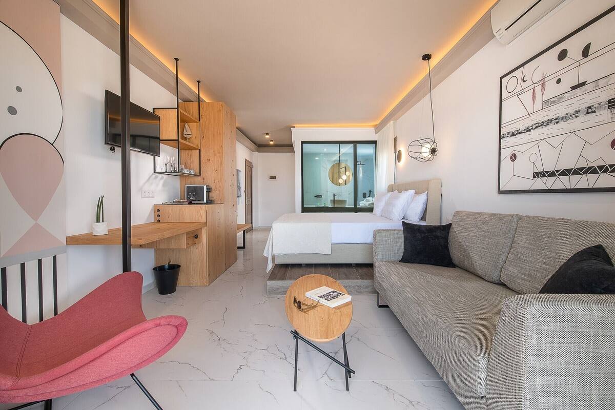 A modern hotel room with a bed, a sofa, a red chair, a small wooden table, a TV, and a large window.