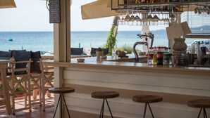 A seaside bar with a menu board listing various coffee and espresso drinks, a bar counter with bottles and glasses, and outdoor seating with a view of the ocean. - Assano Sea Front Luxury (Sithonia)