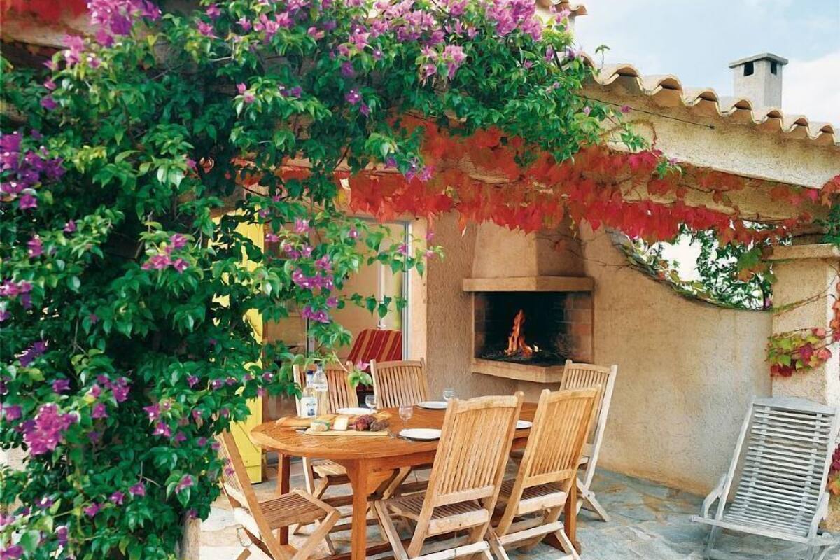Eine Terrasse mit einem runden Holztisch und Stühlen, einem Backofen aus Ziegeln und einer Pergola mit blühenden Weinreben.