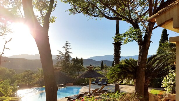 A pool area with lounge chairs, a thatched-roof structure, and a view of mountains. - Les Toits de Santa Giulia (Porto-Vecchio)