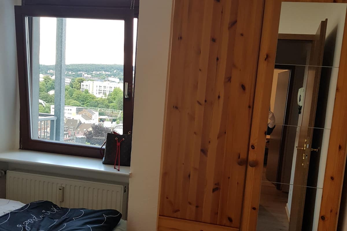 A bedroom with a large window, wooden wardrobe, and a bed with a black and white patterned bedspread.