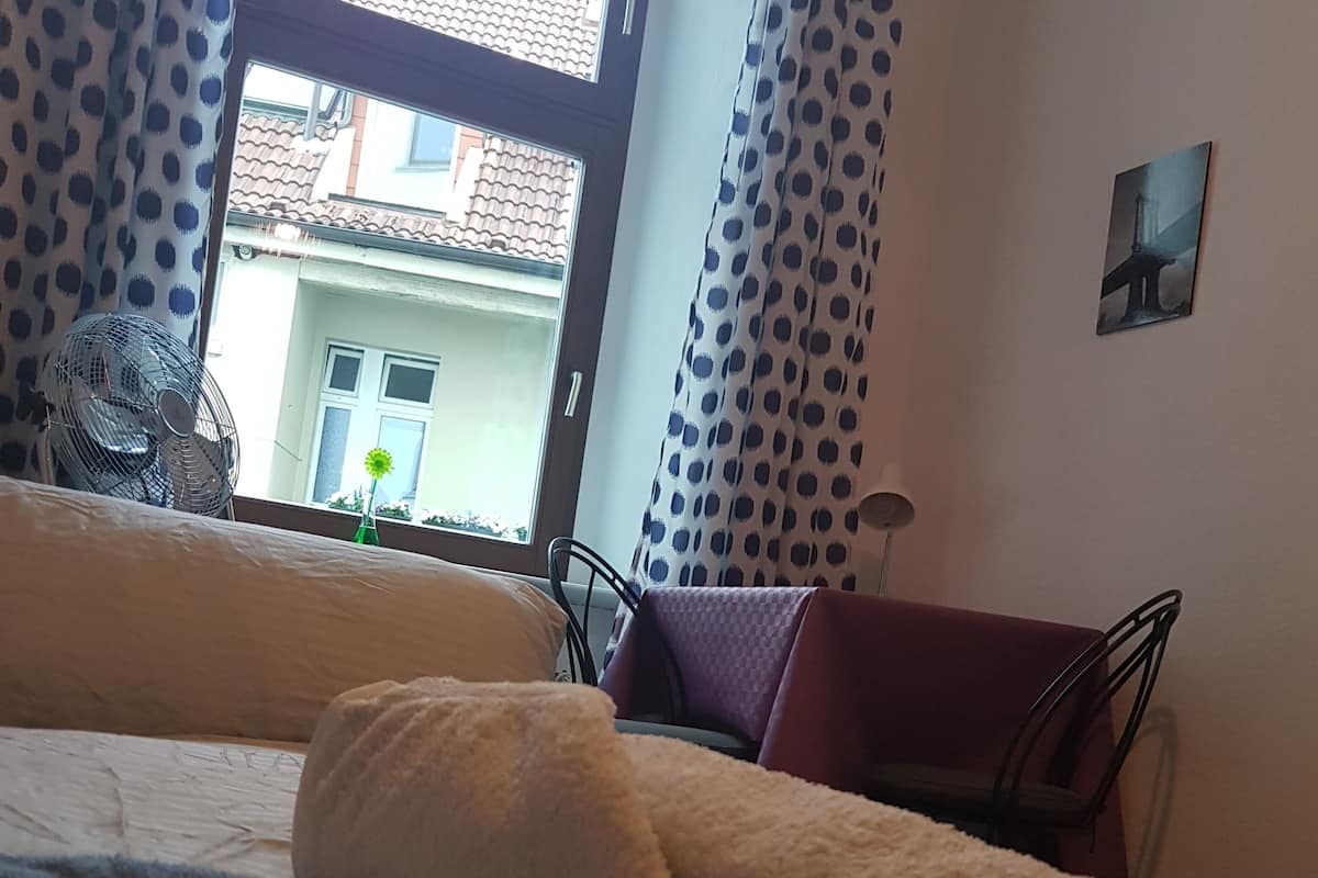 A bedroom with a bed, a window with curtains, a fan, and a view of buildings outside.