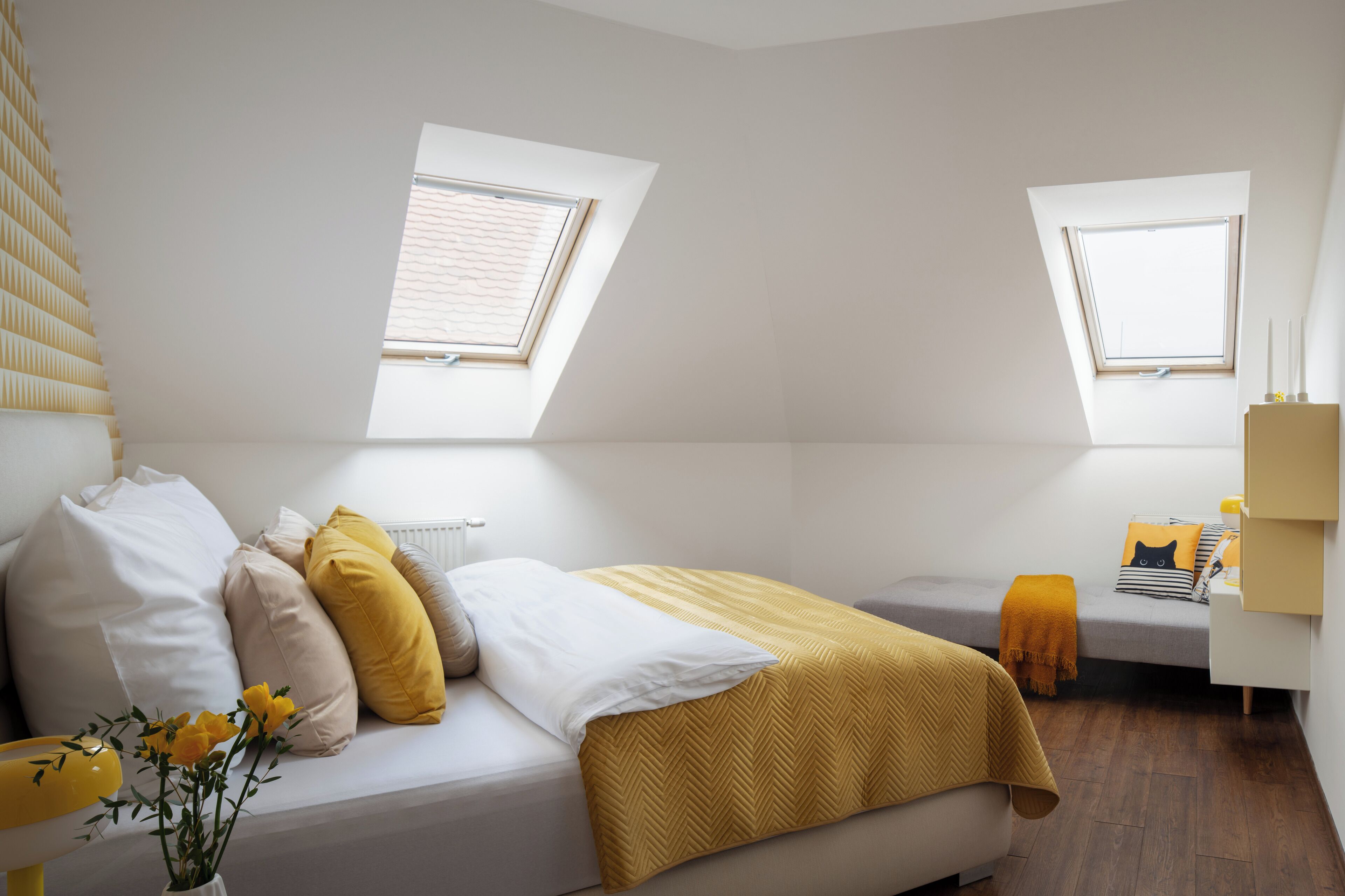 A bedroom with a skylight, a bed with white and yellow bedding, a bedside table, and a small bedside table with a lamp.