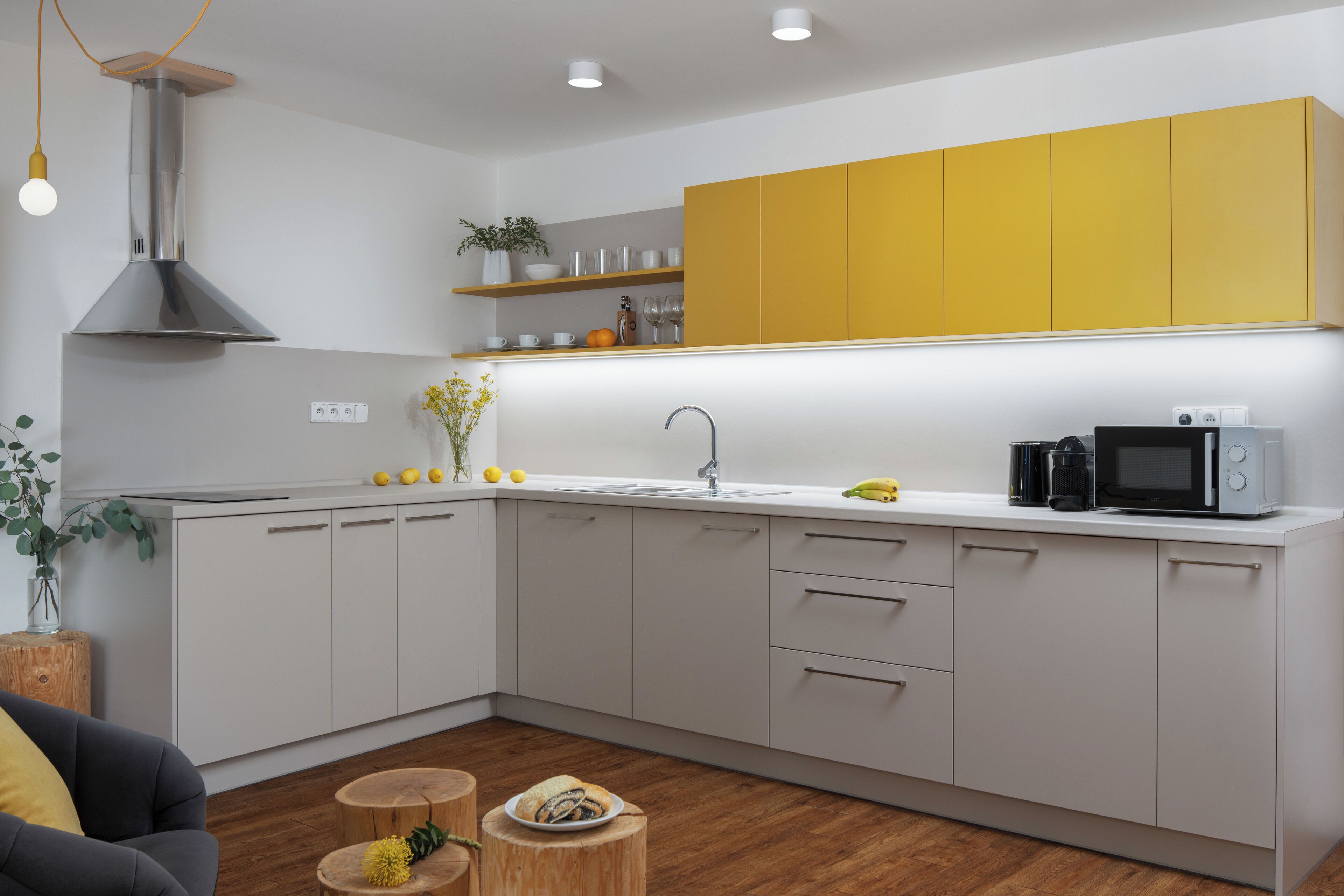 A modern kitchen with white cabinets, a yellow upper cabinet, a stainless steel range hood, a microwave, and a sink.
