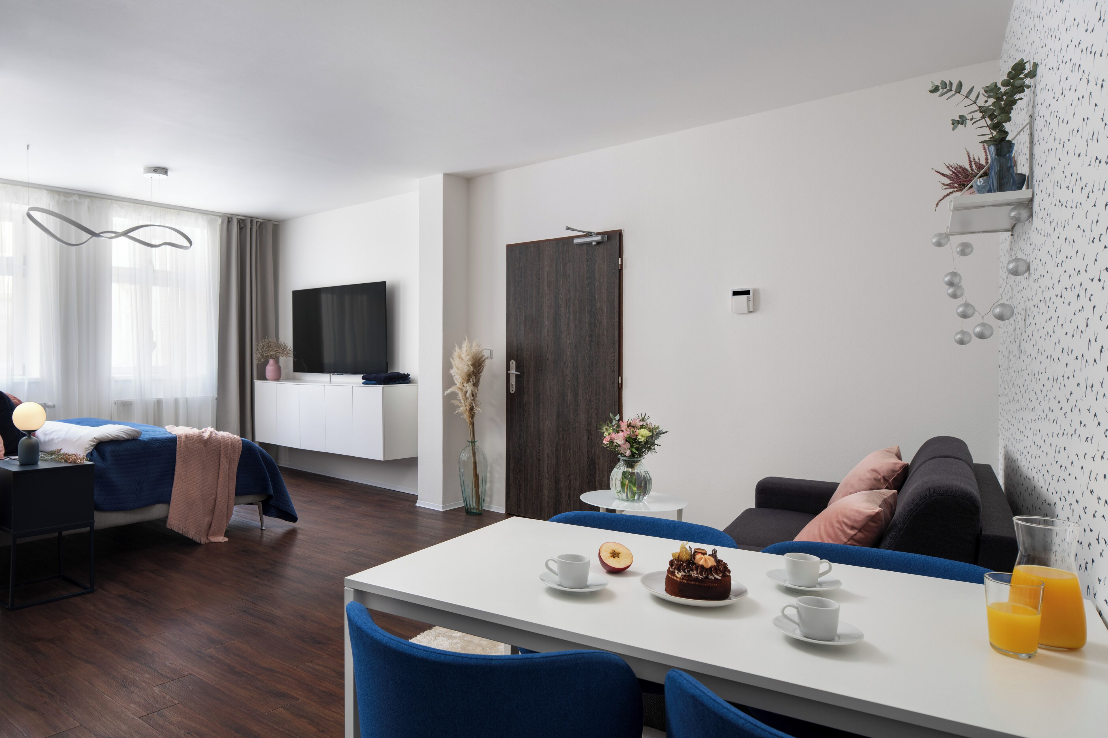 Ein modernes Hotelzimmer mit einem Bett, einer Essecke mit Tisch und Stühlen, einem an der Wand montierten Fernseher und einem kleinen Beistelltisch mit Blumen.
