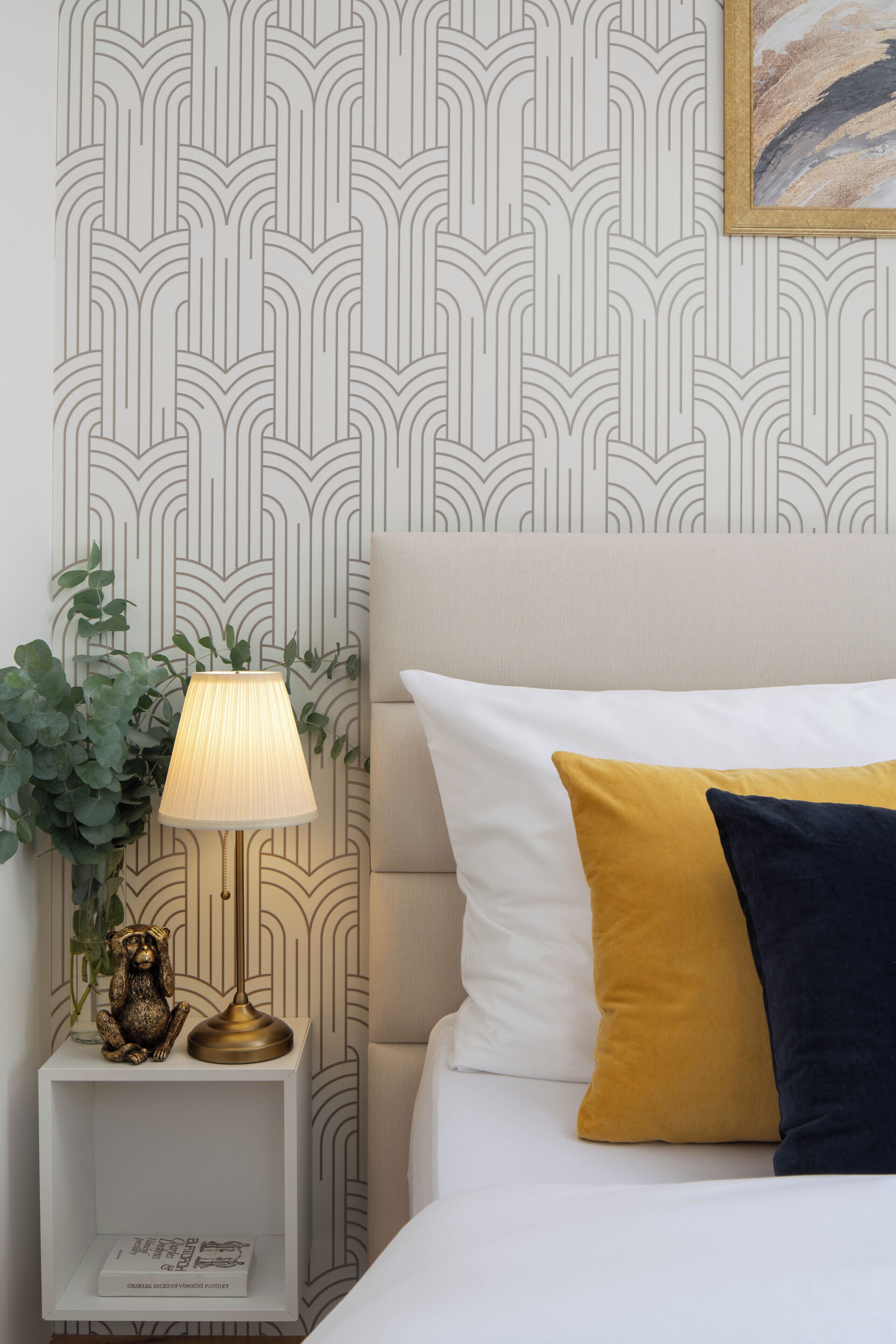 A neatly arranged bedroom with a patterned wallpaper, a bedside lamp, a small shelf with a decorative item, and a bed with two pillows.