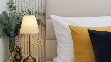 A neatly arranged bedroom with a patterned wallpaper, a bedside lamp, a small shelf with a decorative item, and a bed with two pillows.