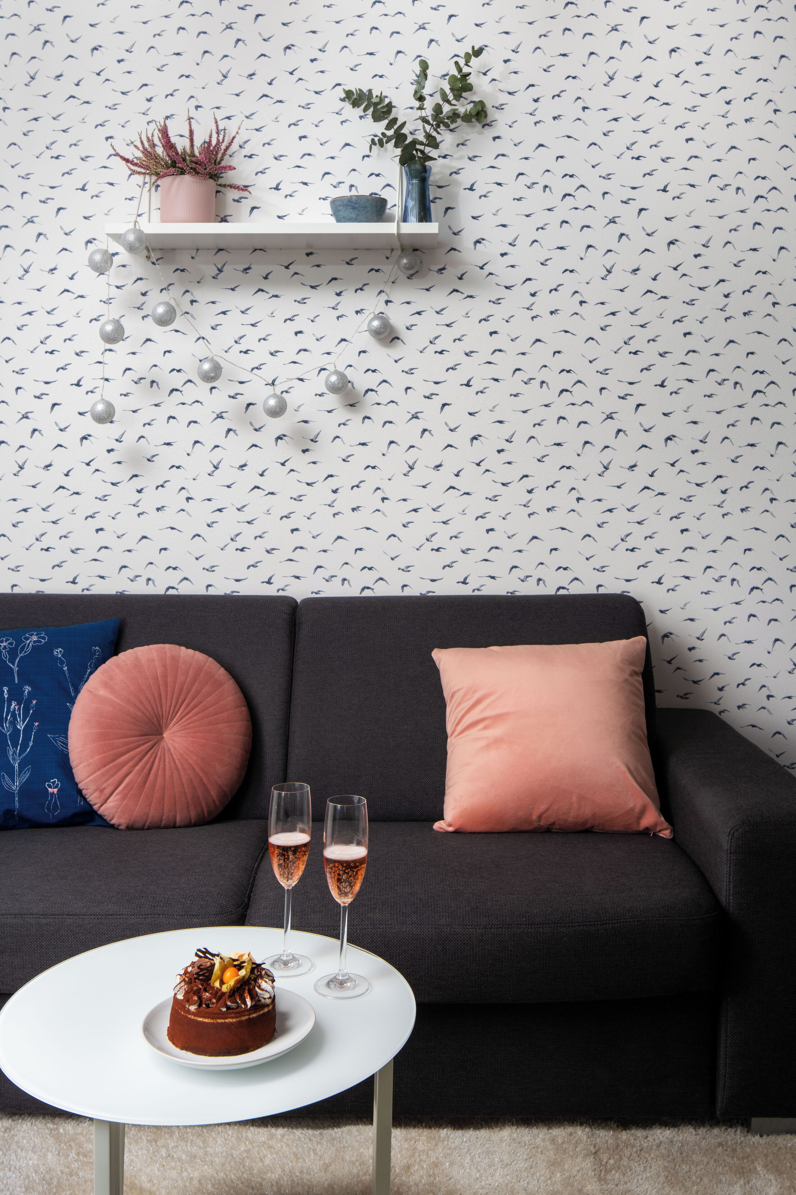 Een moderne woonkamer met een zwarte bank, twee kussens, een witte salontafel met een taart en twee champagneglazen, en een decoratieve plank met planten.