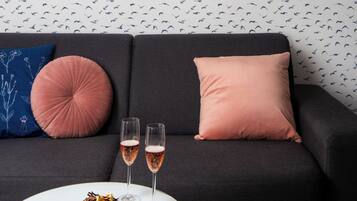 Een moderne woonkamer met een zwarte bank, twee kussens, een witte salontafel met een taart en twee champagneglazen, en een decoratieve plank met planten.