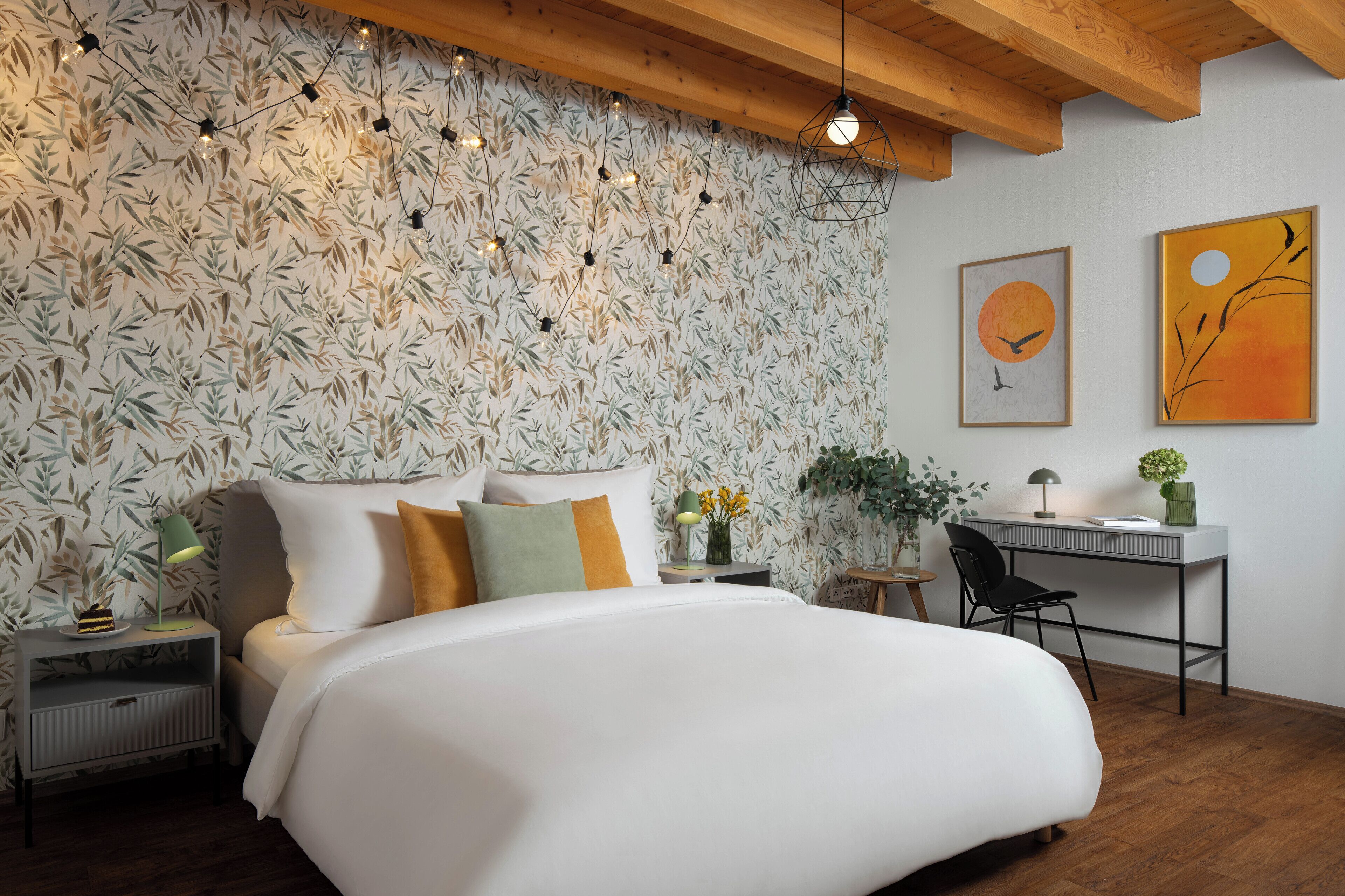 A bedroom with a large bed, a desk, and a chair. There is a wall with a leafy wallpaper and two framed artworks.