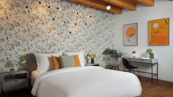 A bedroom with a large bed, a desk, and a chair. There is a wall with a leafy wallpaper and two framed artworks.