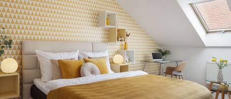 A modern bedroom with a geometric patterned wall, a bed with a gold throw, a desk with a laptop, and a skylight.