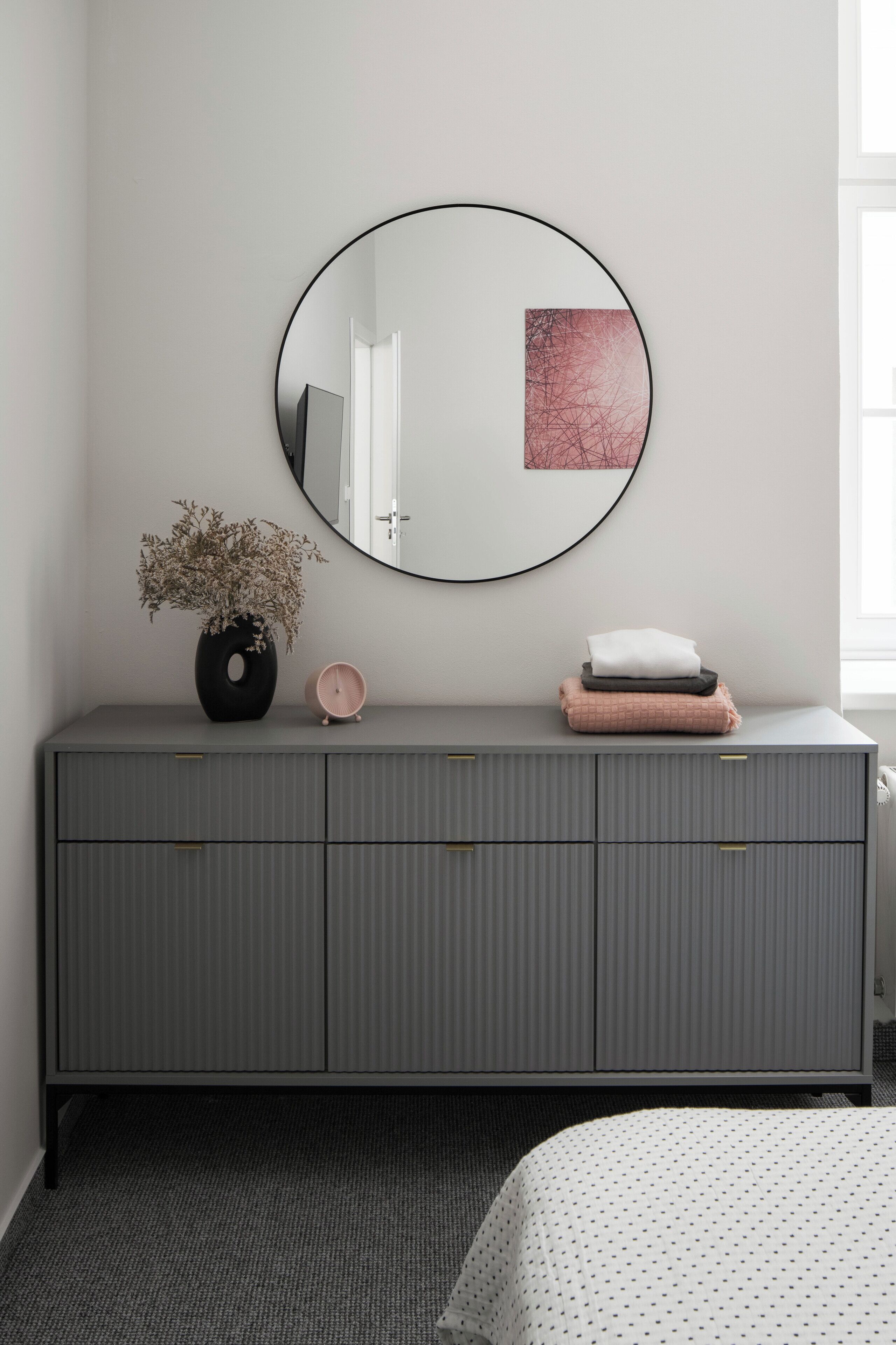 Une commode grise avec un miroir rond, un vase contenant des fleurs séchées, une horloge rose et des serviettes pliées.