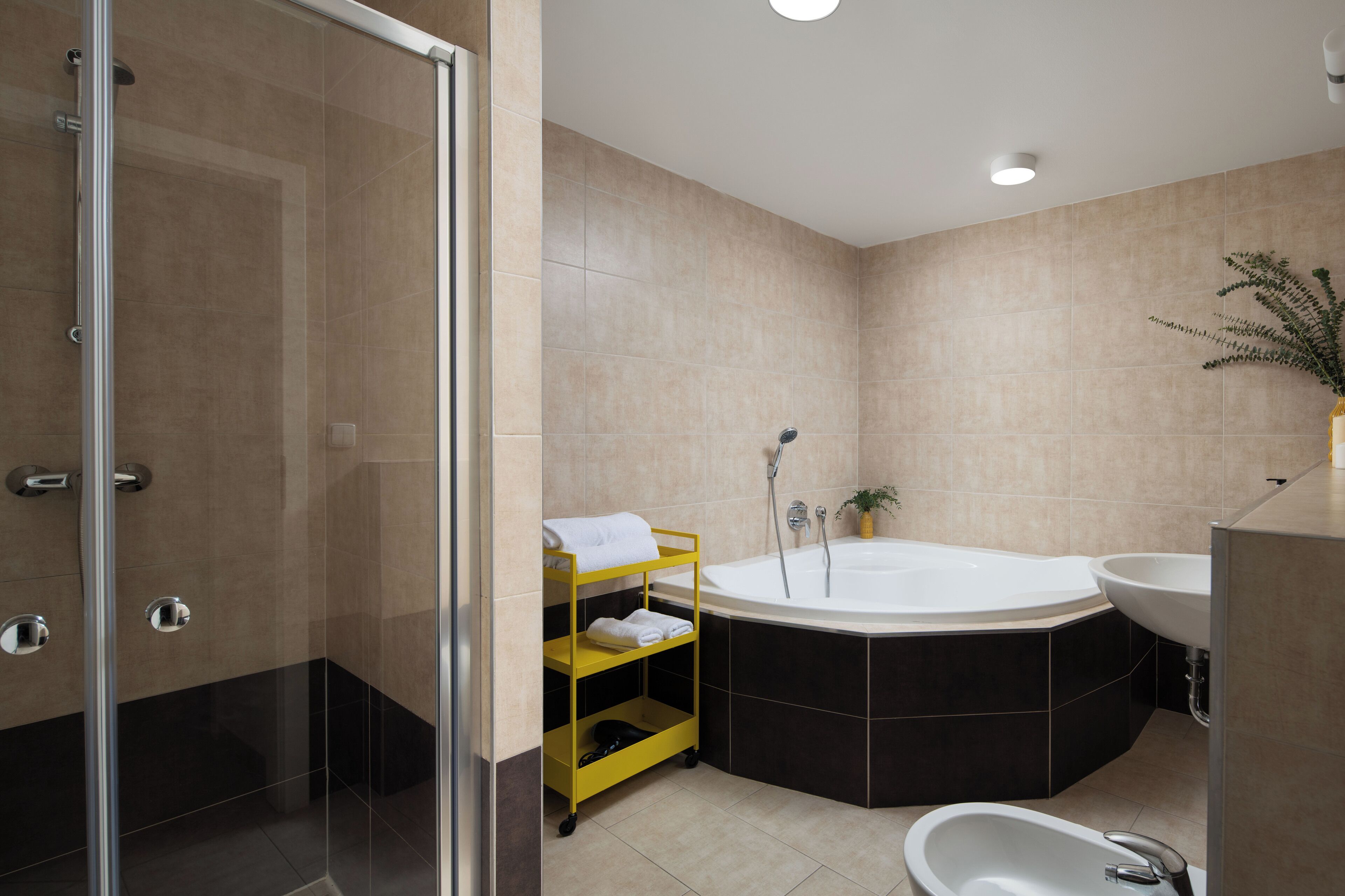 A modern bathroom with a glass-enclosed shower, a freestanding bathtub, and a yellow storage cart.