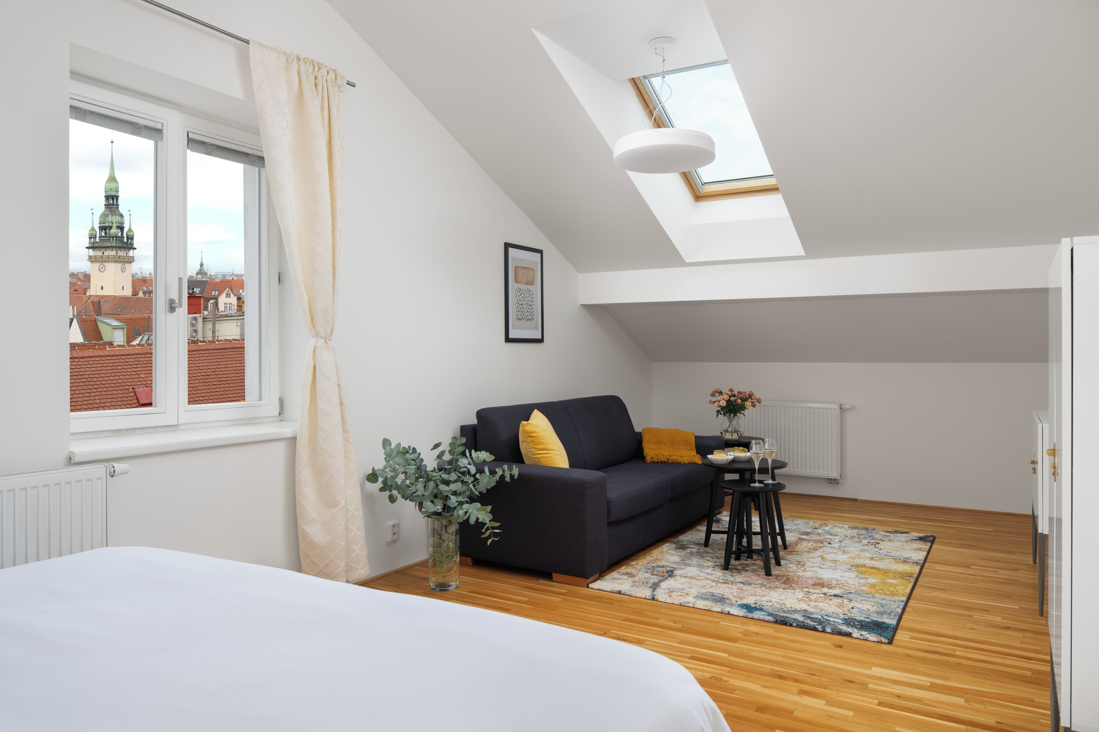 A modern room with a skylight, a bed, a sofa, a small table with a vase, and a view of a cityscape.