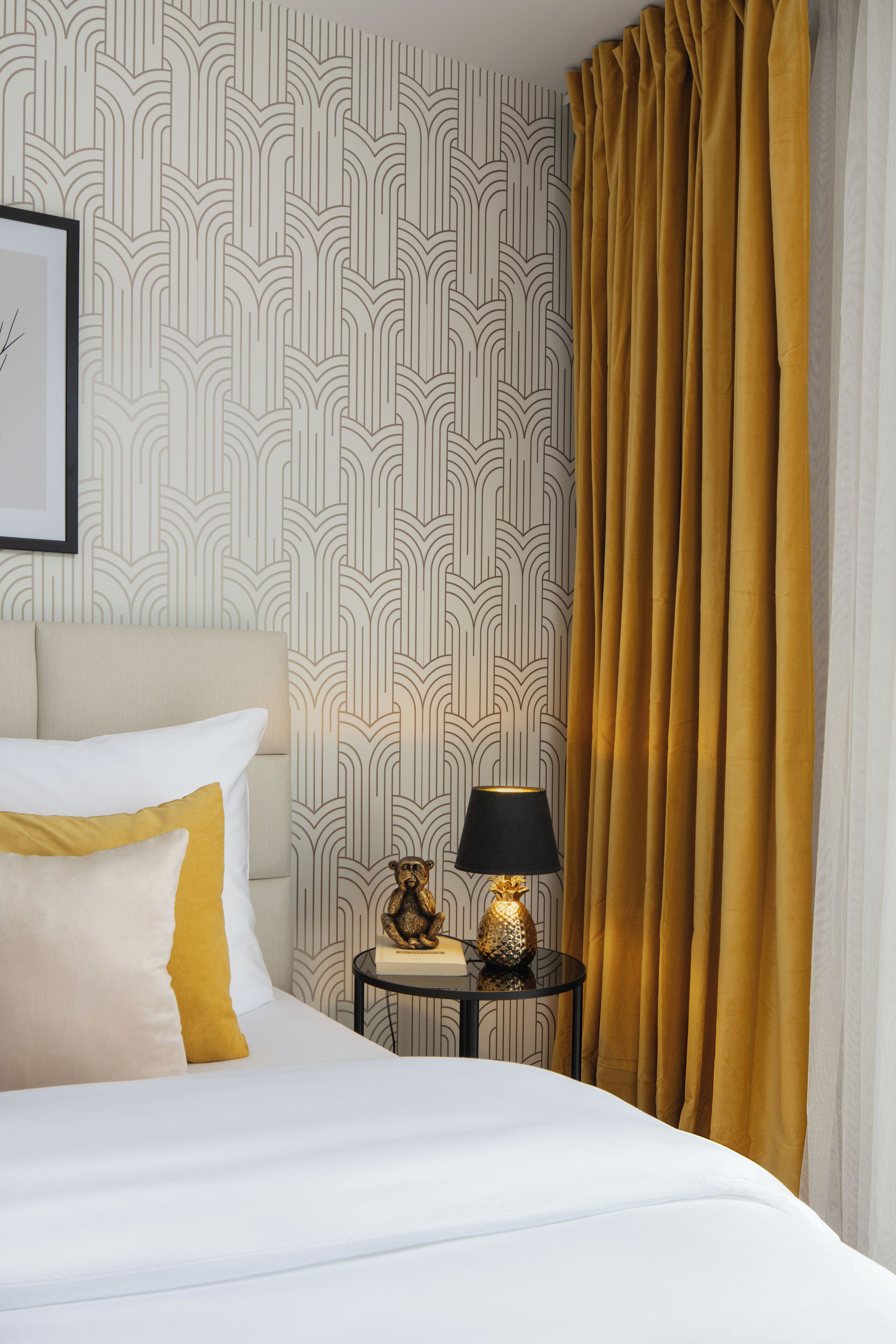A neatly made bed with white and mustard pillows, a small round table with a lamp, and a gold decorative item.