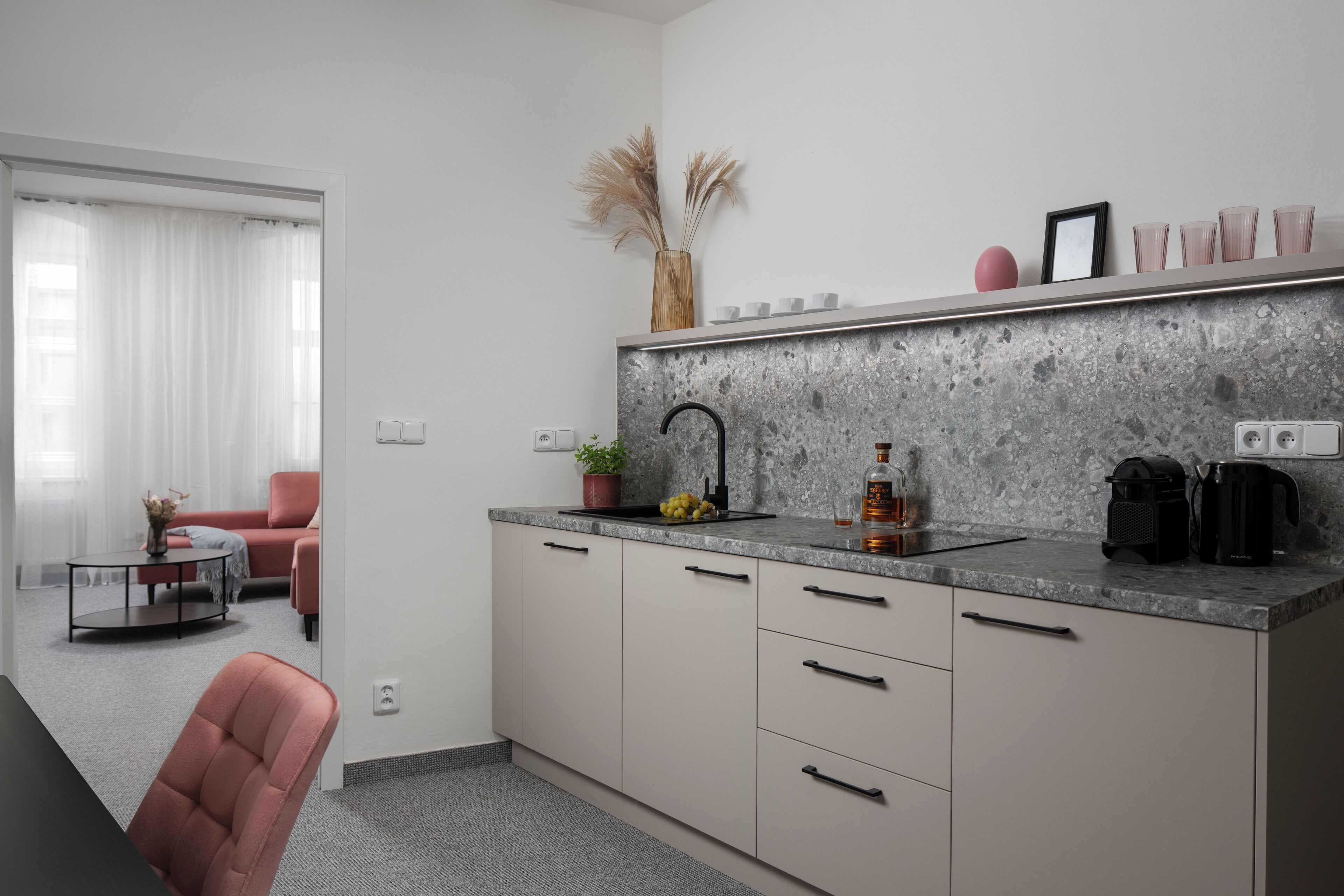 A modern kitchen with a granite countertop, a sink, and a coffee machine. There is a dining area with a table and chairs visible through an open door.