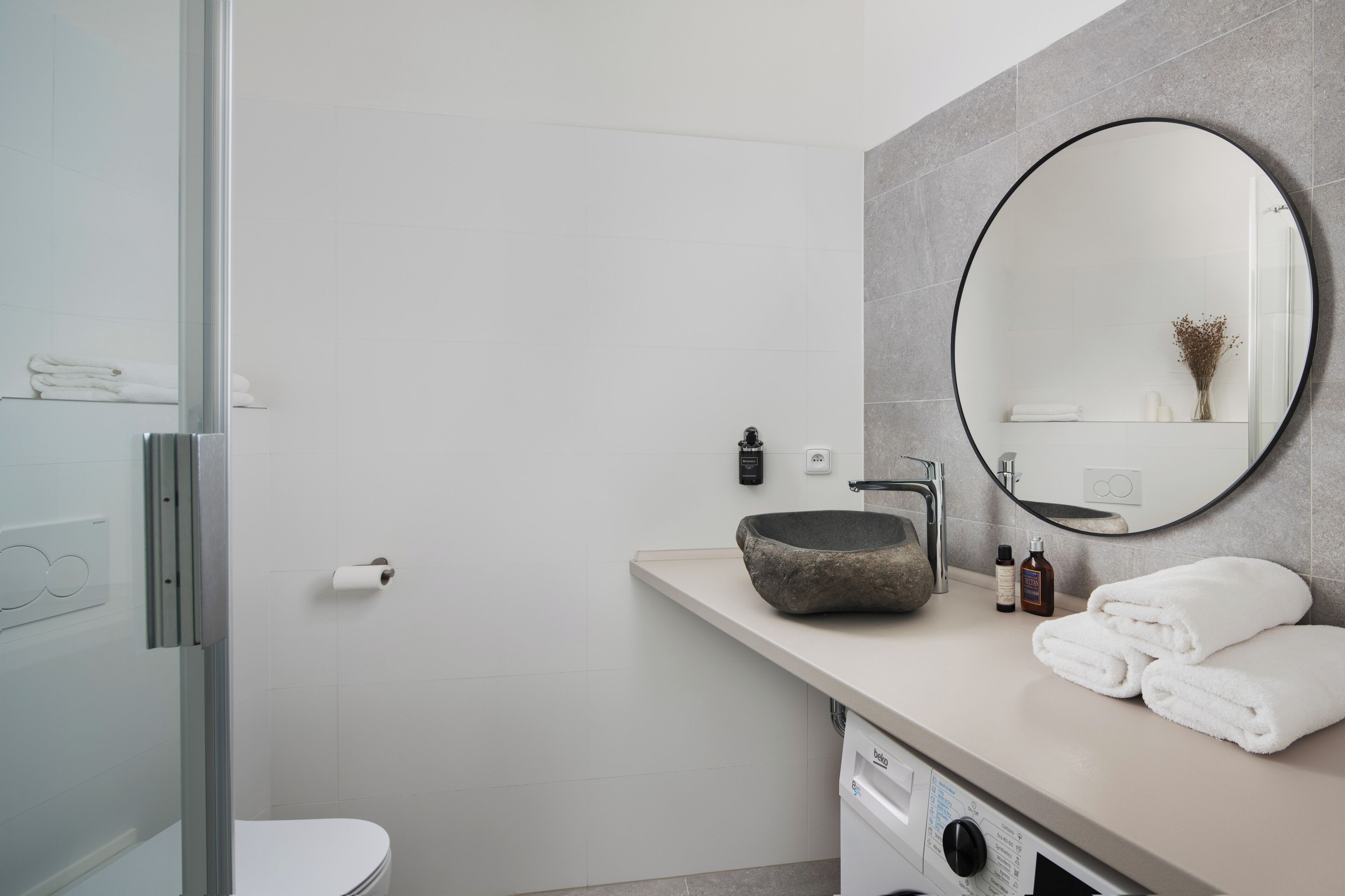 Un baño moderno con cabina de ducha de vidrio, un lavabo color crema con lavamanos de piedra, un espejo redondo y toallas dobladas con esmero.