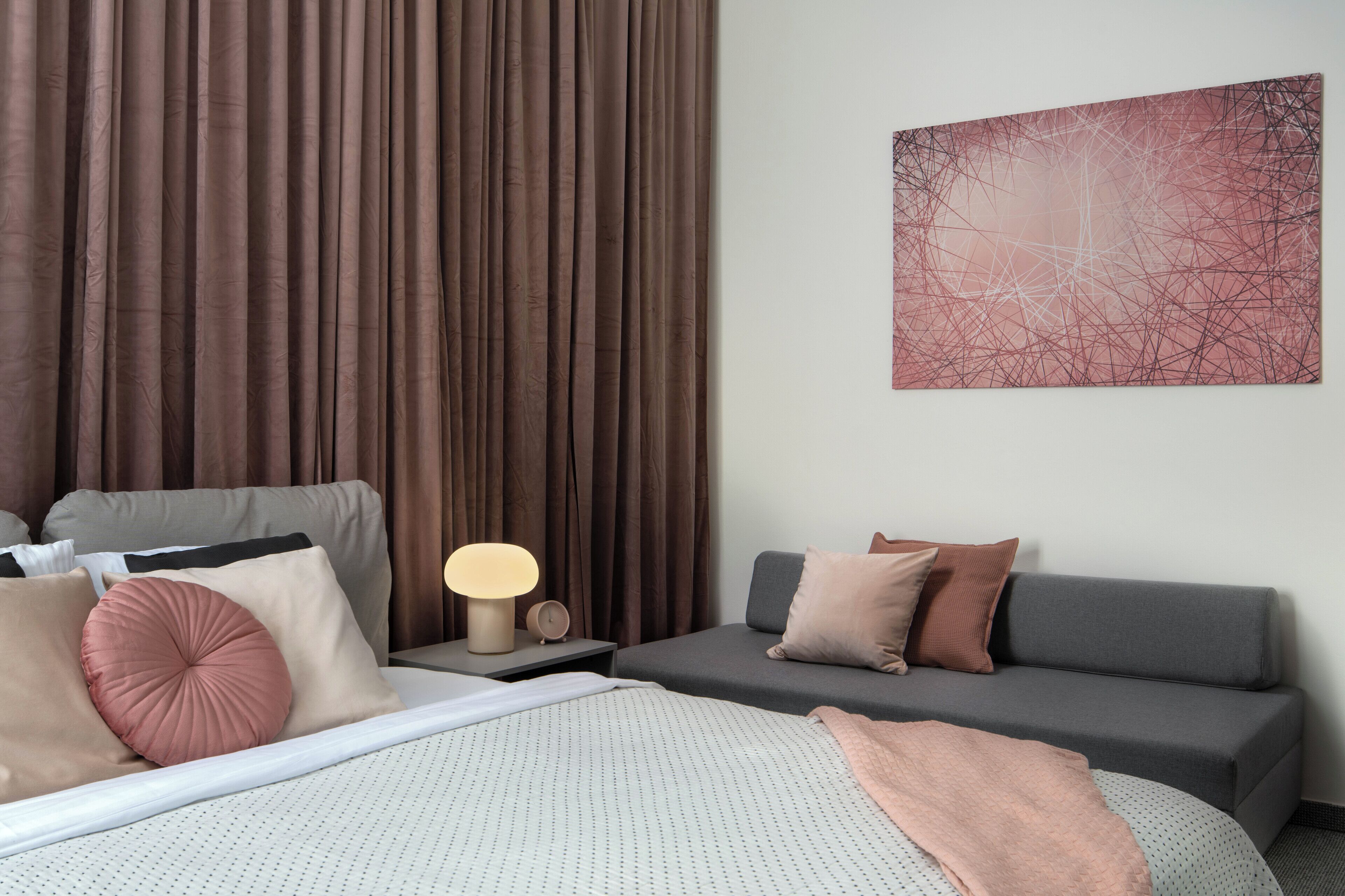 A modern hotel room with a bed, a sofa, a nightstand with a lamp, and a painting on the wall.