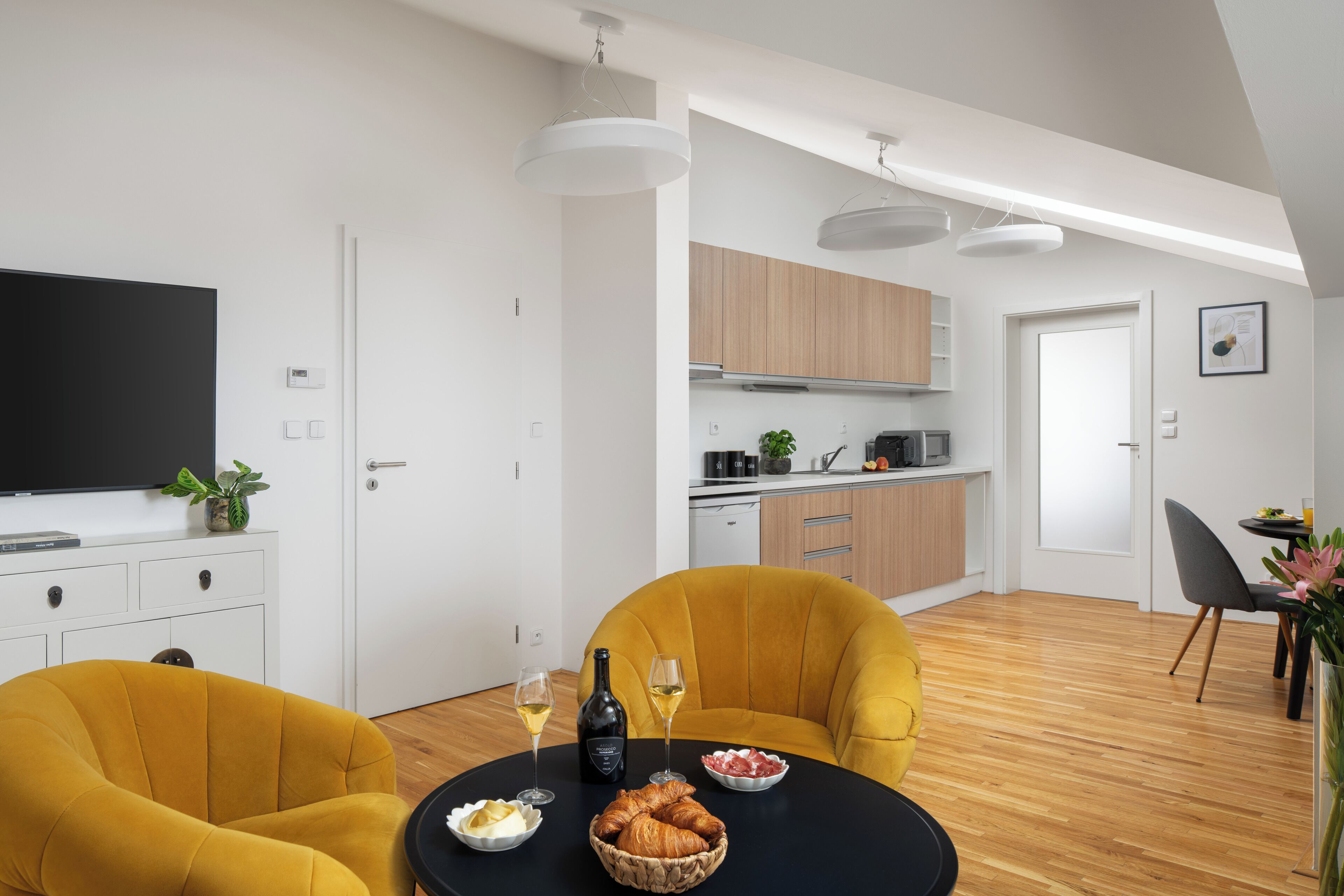 Un salon moderne avec un téléviseur noir, des fauteuils jaunes, une table à manger avec de la nourriture et des boissons, et une cuisine en arrière-plan.