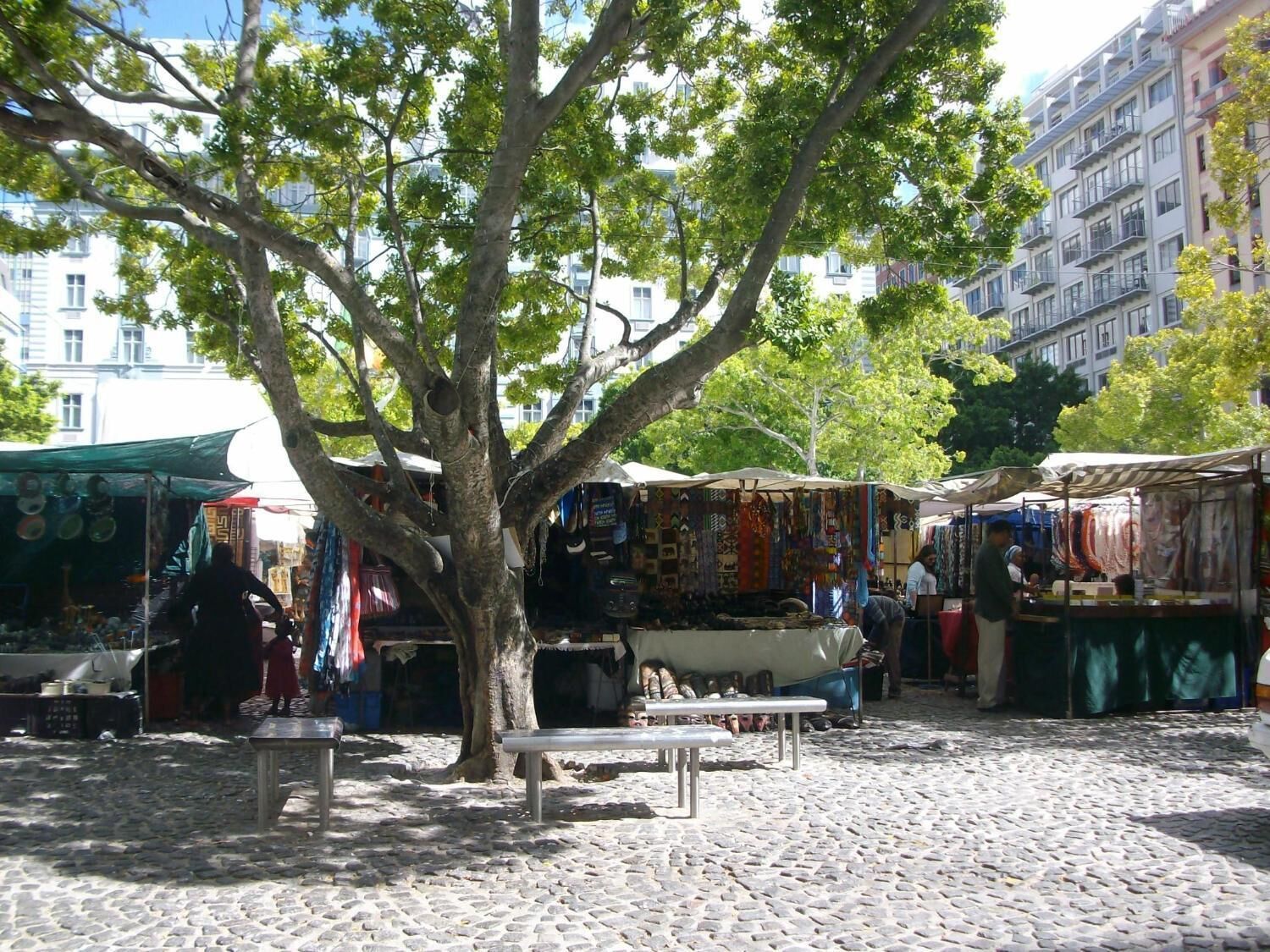 Une rue pavée bordée d’étals de marché et ombragée par un arbre.