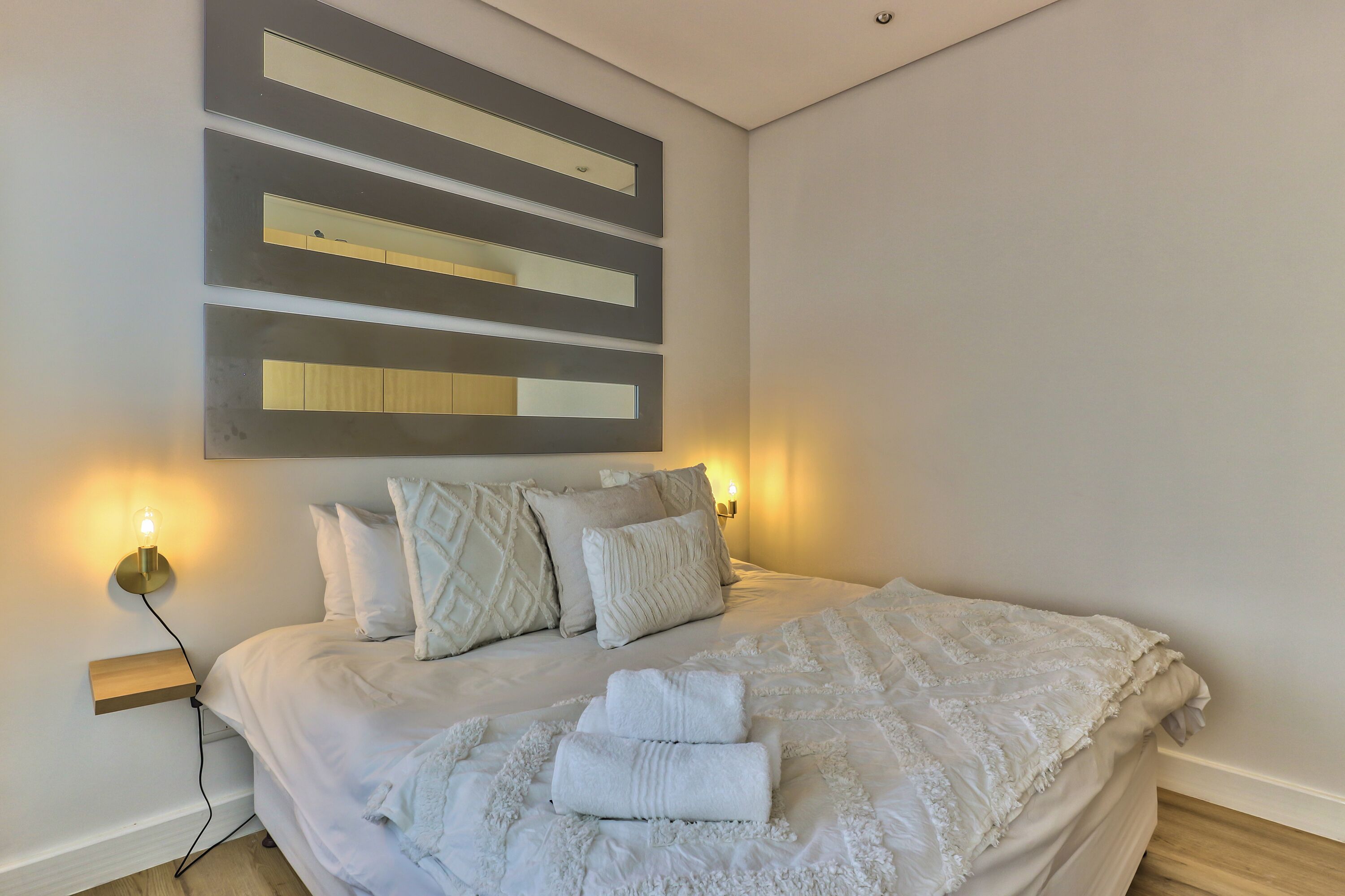 A neatly made bed with white linens and a folded towel, a bedside table with a lamp, and a wall-mounted light fixture.
