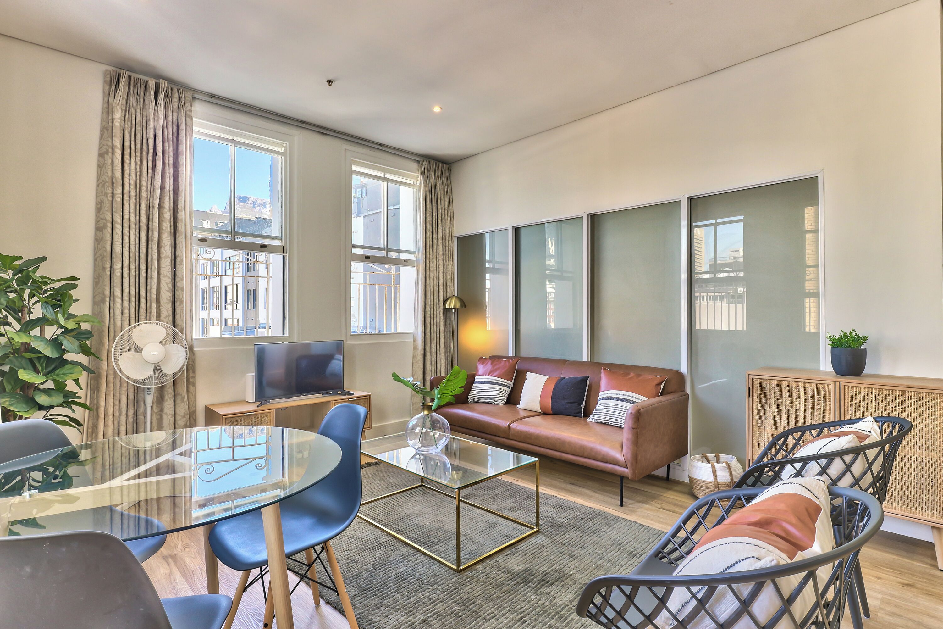A modern living room with a glass coffee table, blue chairs, a leather sofa, and a large window with curtains.
