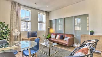 A modern living room with a glass coffee table, blue chairs, a leather sofa, and a large window with curtains.