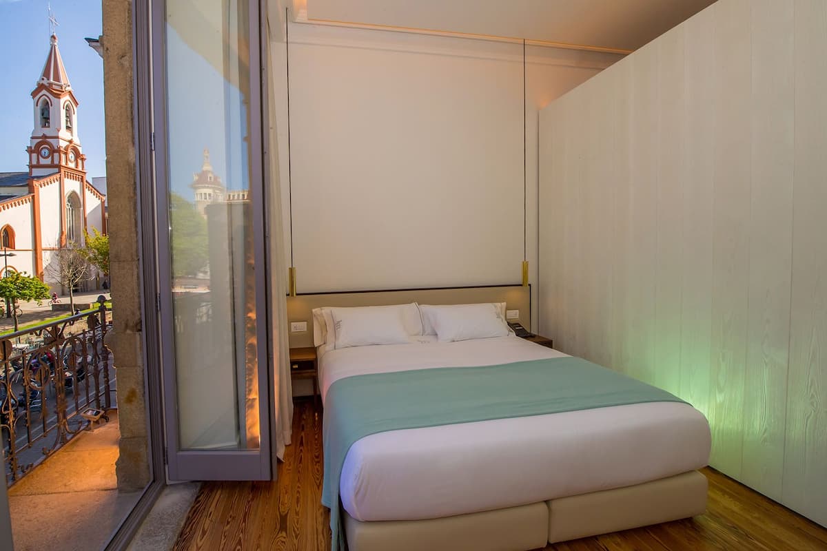 A hotel room with a bed, a nightstand, and a view of a church through a glass door.