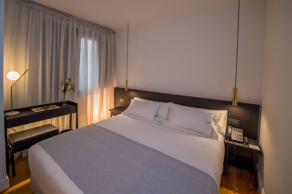 A modern hotel room with a large bed, a desk, a chair, a bedside table with a phone, and a lamp.
