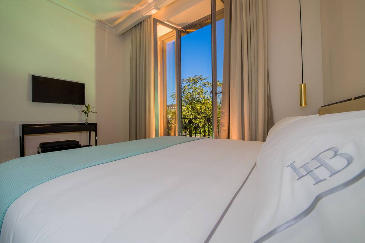 A hotel room with a large bed, a flat-screen TV, a bedside table, and a view of greenery through the window.