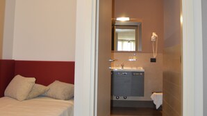 Un couloir étroit mène à une salle de bain équipée d’un lavabo et d’un miroir. On aperçoit un lit avec une tête de lit rouge sur la gauche.