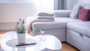Una sala moderna con un sofá blanco, una mesita redonda con una planta en maceta y un control remoto, además de un ventanal con cortinas.
