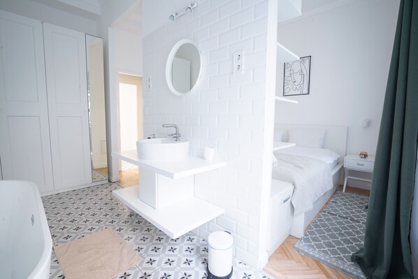 A modern, white-themed bedroom with a large bed, a bedside table, a built-in sink, and a mirror.