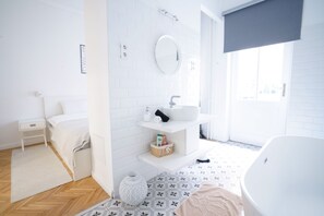 A modern bathroom with a white sink, mirror, and bathtub. There is a small bedside table and a decorative vase on the floor.