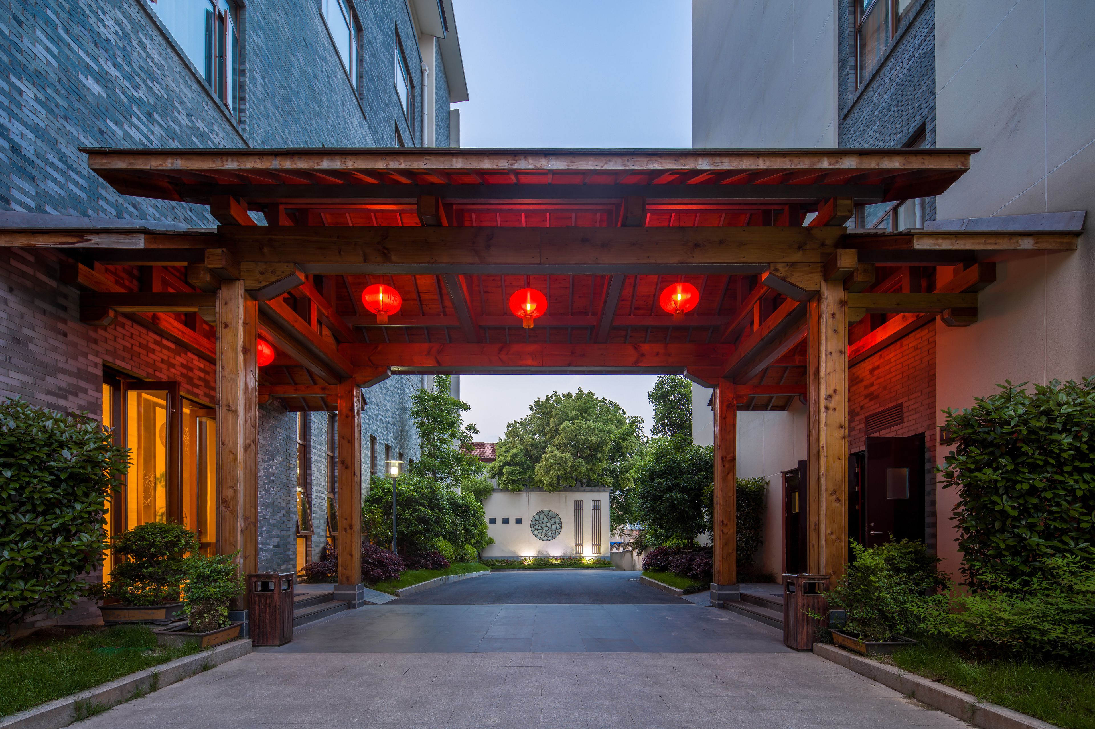 A covered entrance with a wooden structure and red lanterns, leading to a building with a circular window and a brick wall.