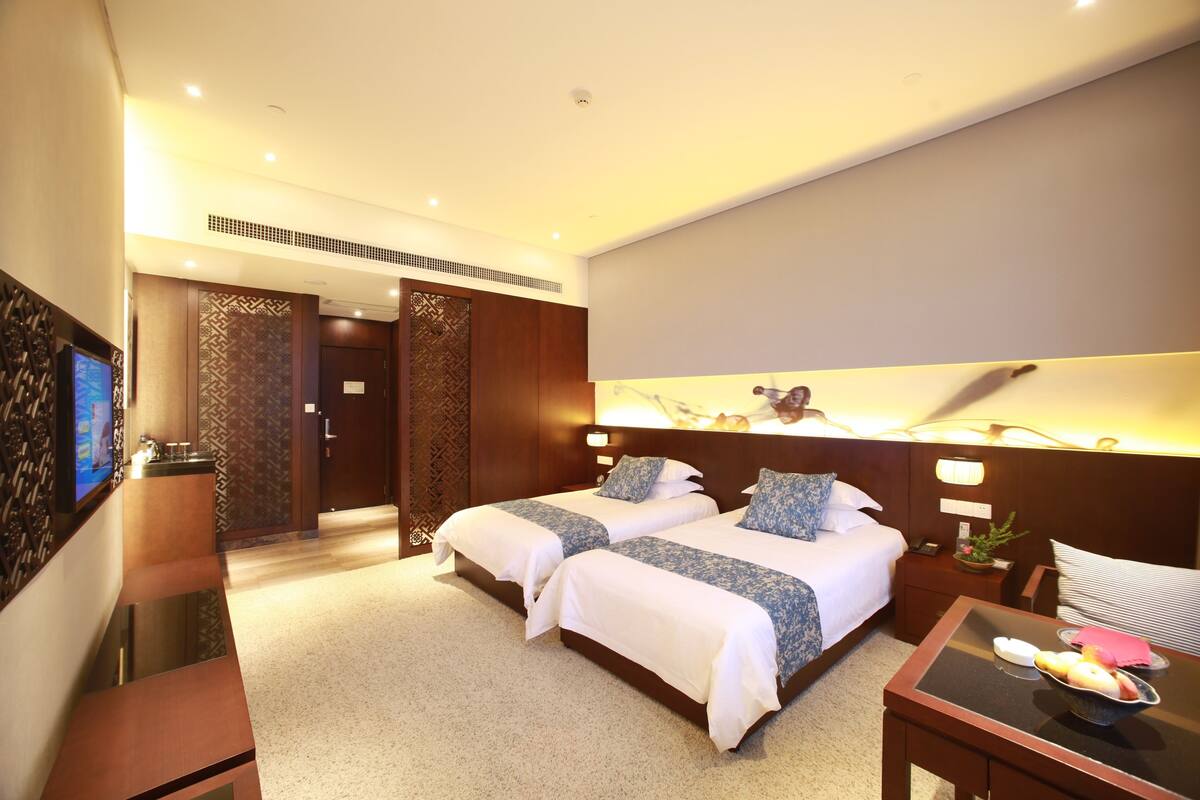 Habitación de hotel con dos camas, un escritorio, un televisor y un baño visible a través de una puerta abierta.