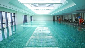 A large indoor swimming pool with clear glass walls and a skylight, surrounded by lounge chairs and life-saving equipment. - Grand New Century Hotel Yixing (Wuxi)