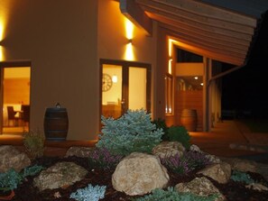 A modern house with a well-lit entrance, a large barrel, and a garden with rocks and plants. - Agriturismo Al Maniero (Trento)