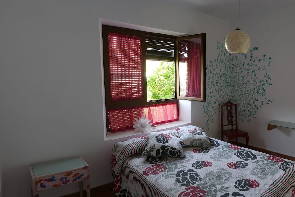 A bedroom with a bed, a window with blinds, a chair, a shelf with a vase, and a mirror with a decorative frame.