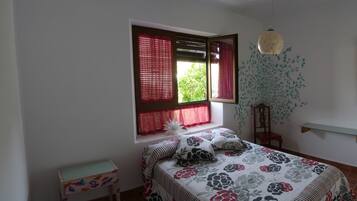 Un dormitorio con cama, una ventana con persianas, una silla, una estantería con un jarrón y un espejo con marco decorativo.