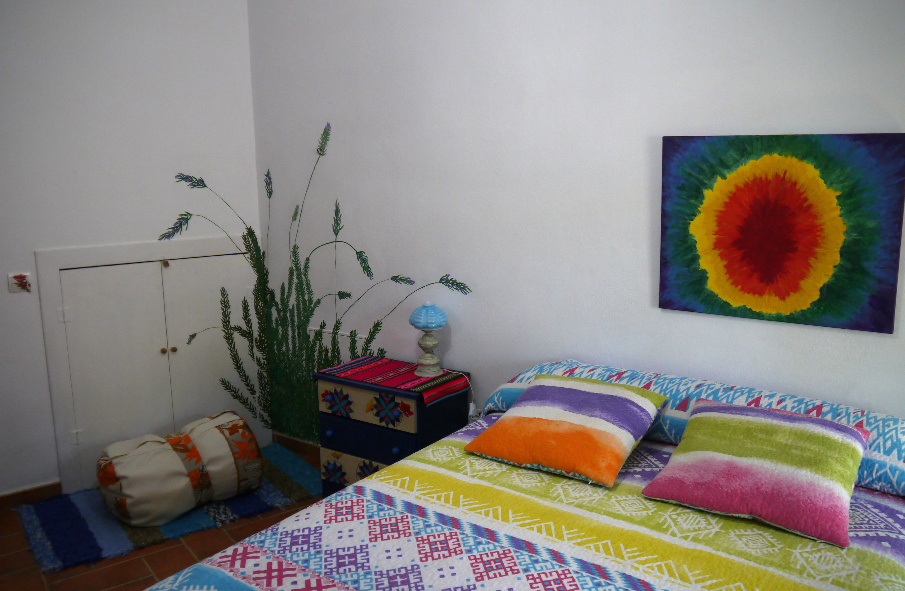 Un dormitorio con cama, almohadas coloridas, una planta y un cuadro en la pared.