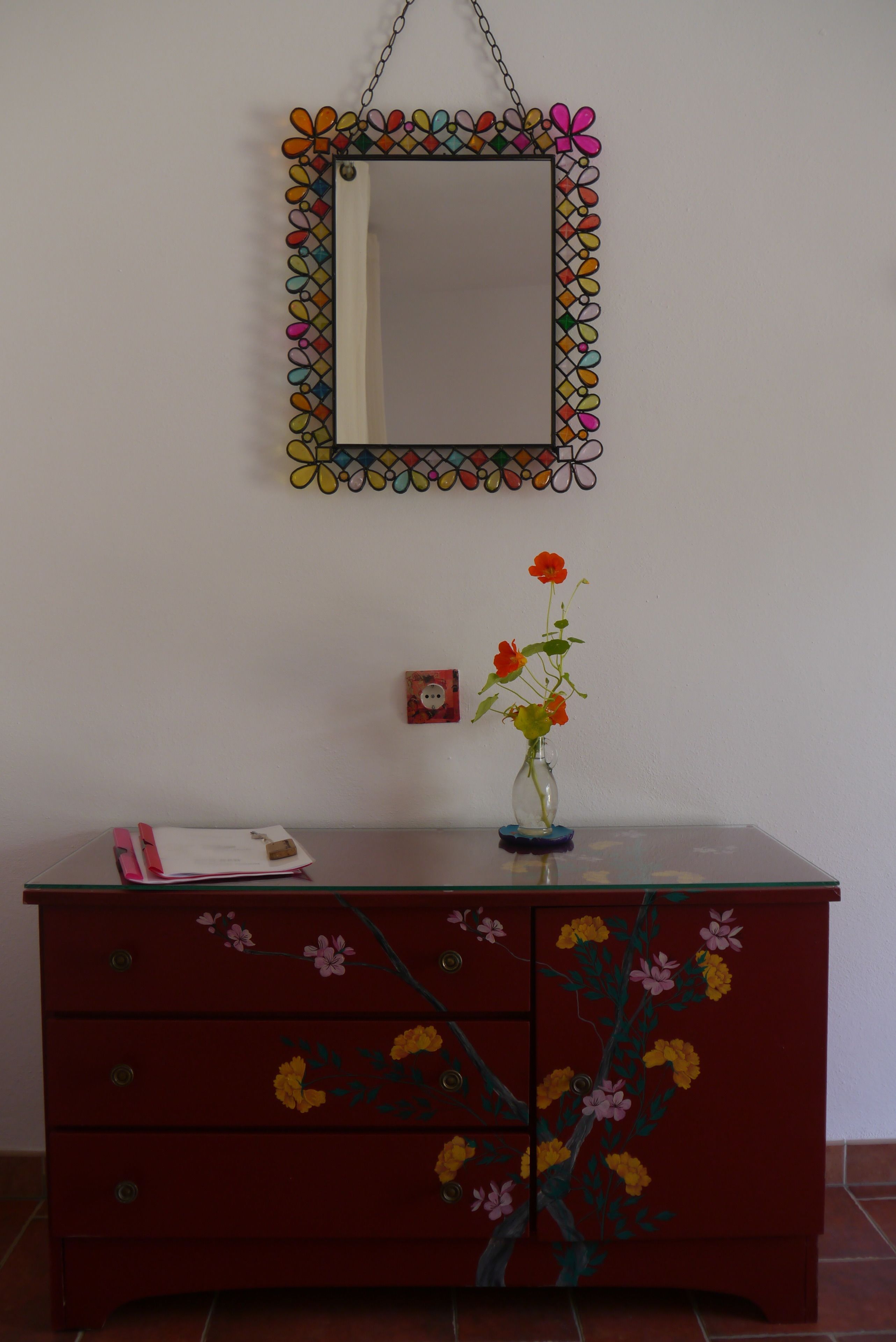 A red dresser with floral designs, a mirror with a colorful border, and a vase with flowers on a table.