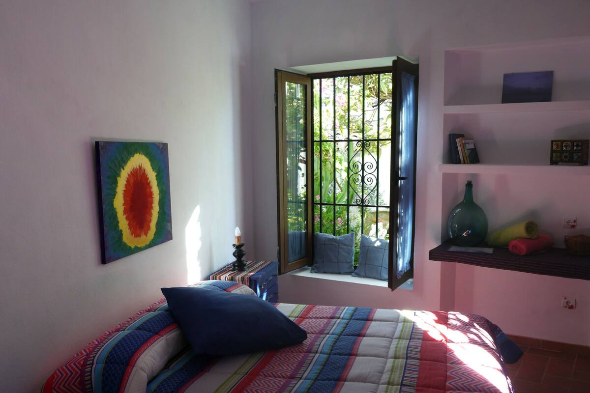A bedroom with a bed, a window with a view of greenery, a shelf with decorative items, and a colorful painting on the wall.