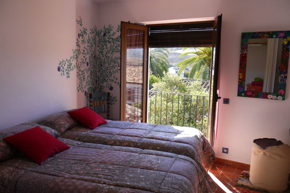 A bedroom with two beds, a balcony with a view of greenery and a house, and a large mirror on the wall.