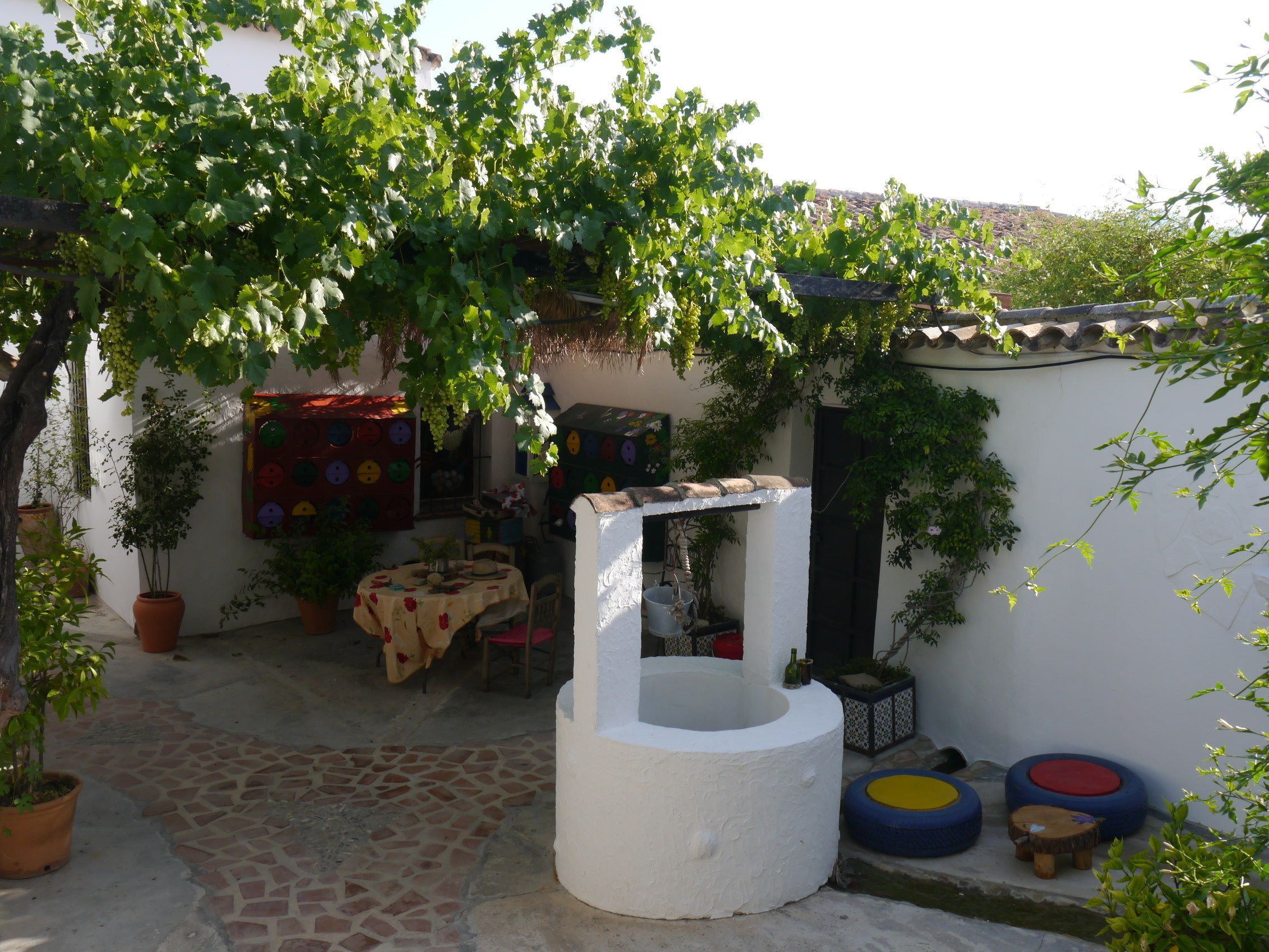 Un patio con una fuente blanca, plantas en macetas, una mesa con sillas y una pieza de arte mural colorida.