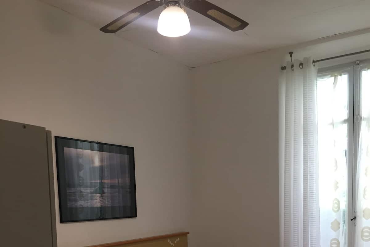 A bedroom with a bed, a nightstand, a wooden dresser, a ceiling fan, and a window with curtains.