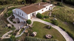A white house with a tiled roof, surrounded by a stone pathway, large rocks, and greenery. - Lu Lisandru (Arzachena)