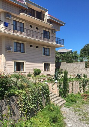 Ein mehrstöckiges Gebäude mit Balkonen, einer Steinmauer und einem Garten mit Stufen, die zum Eingang führen.